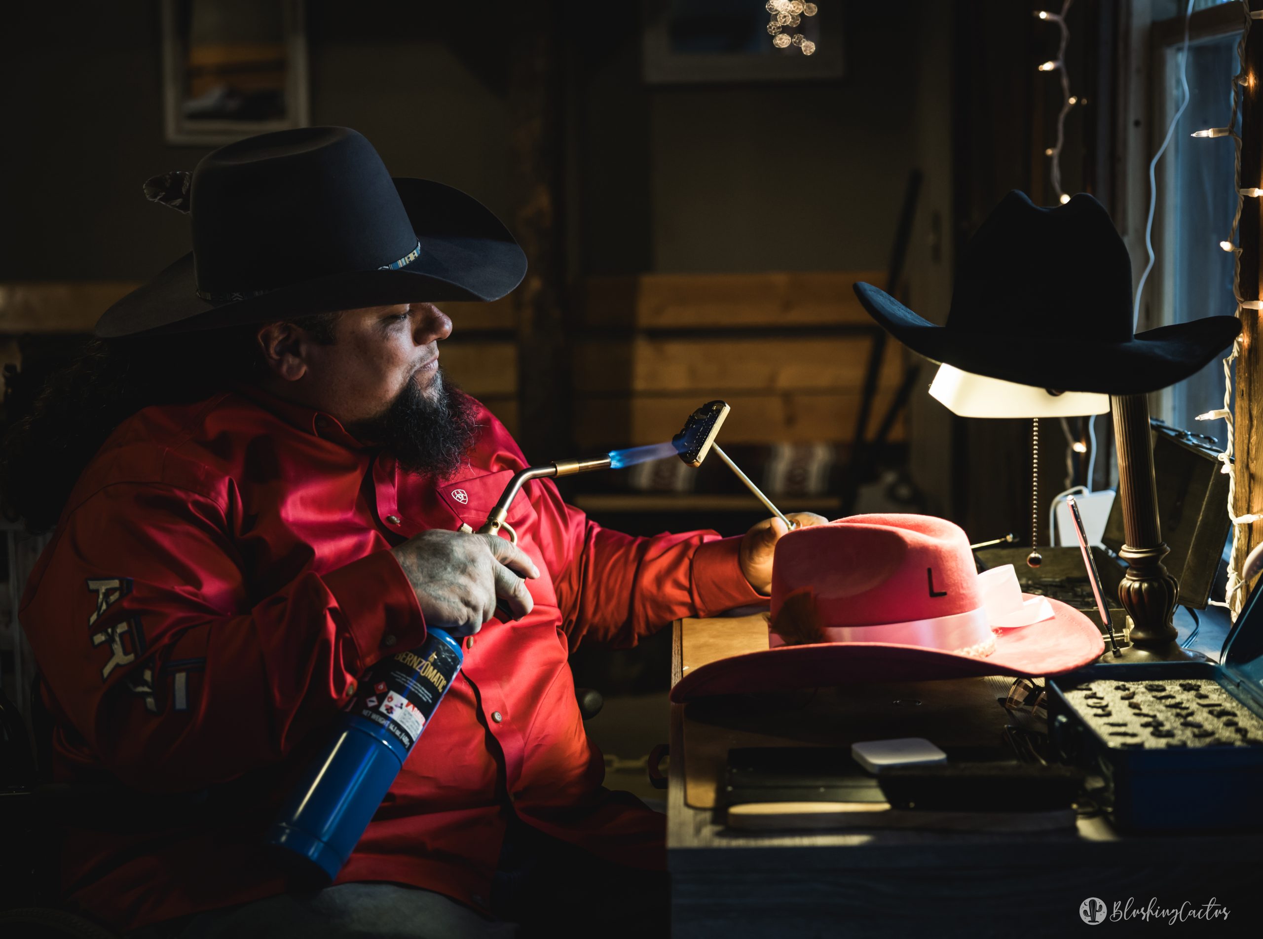 Man shaping hat with flame in workshop, wearing red shirt and black cowboy hat.