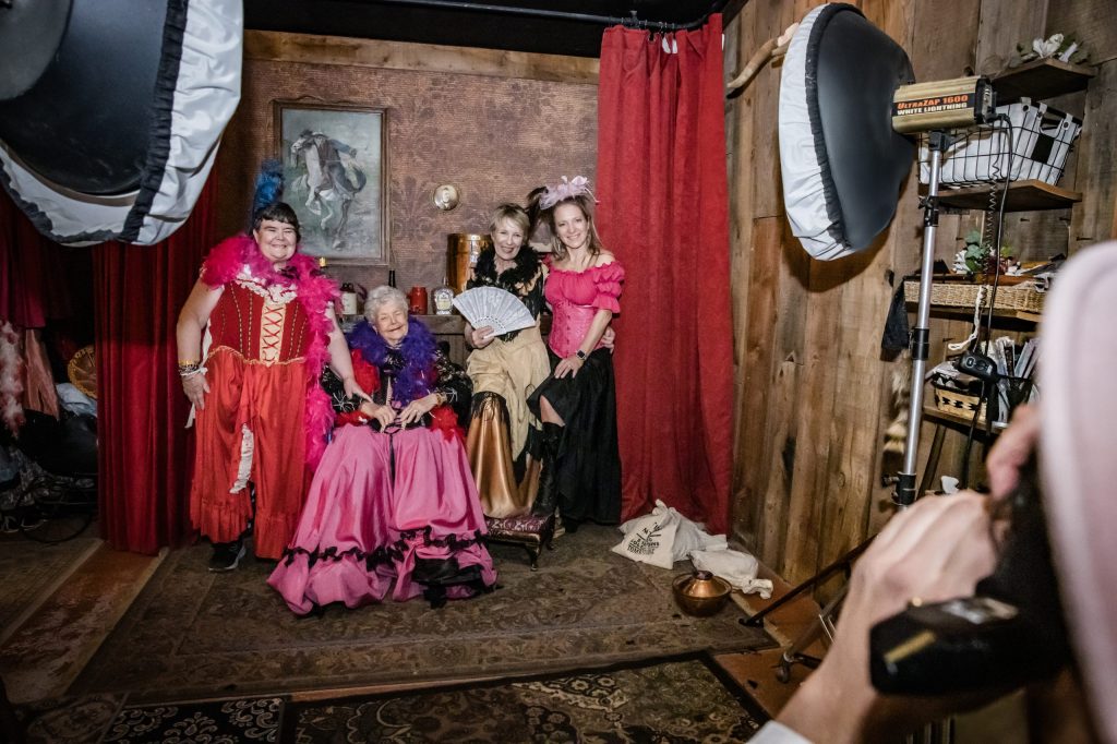 Group in vintage costumes posing indoors with framed portraits on walls.