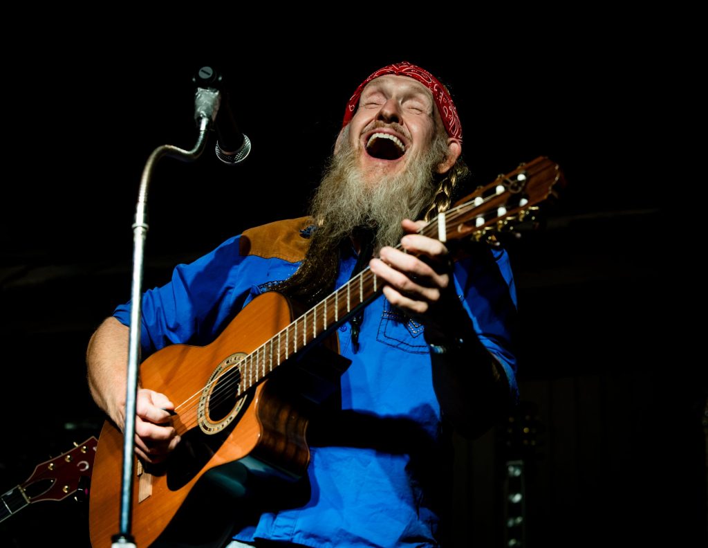 Bearded man with a red bandana plays guitar and sings on stage.