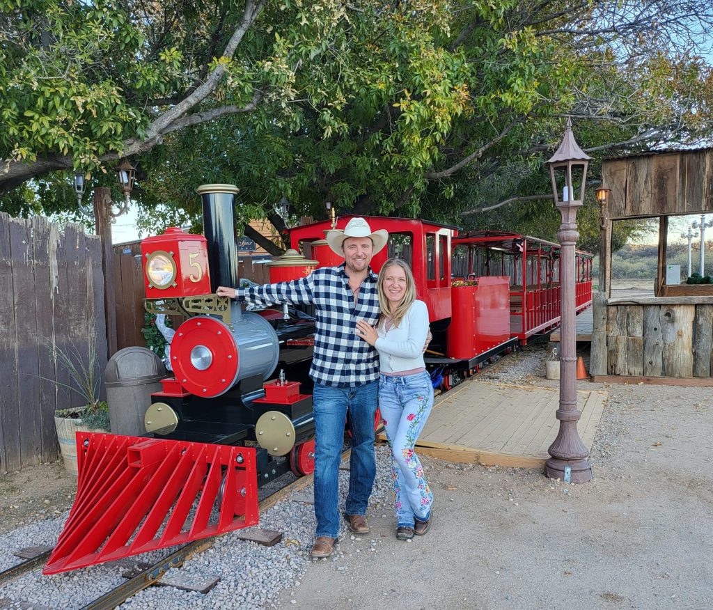 Couple posing in front of a small, stationary red train outdoors.