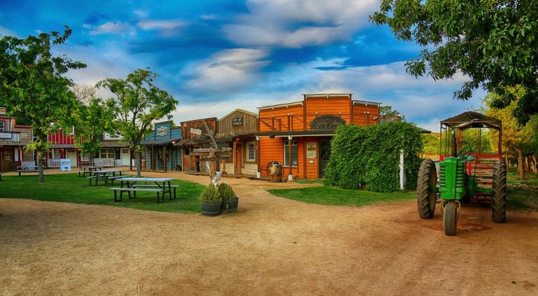 Rustic Western town scene with wooden buildings, tractor, and trees under a blue sky.