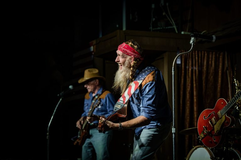 Two musicians in denim shirts and hats perform with guitars on stage.