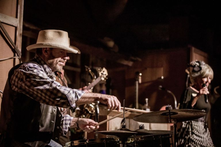 Musician in cowboy hat plays drums beside singer on stage.
