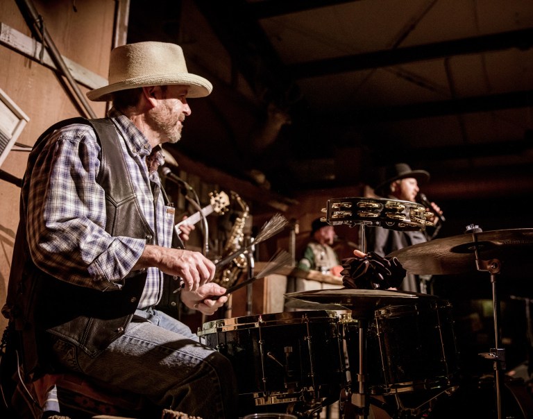 Musicians playing on stage, one with a drum and another singing, both wearing hats.