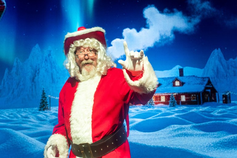 Santa Claus waving with snowy village backdrop and northern lights.